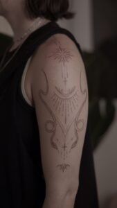 Fine-line ornamental tattoo on the upper arm and shoulder featuring celestial sunbursts, a crescent moon, and symmetrical abstract linework with delicate stippling