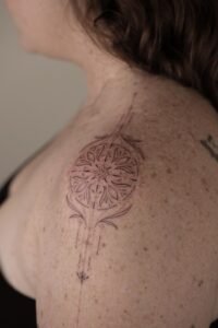 Fine line ornamental mandala tattoo on a freckled shoulder featuring intricate geometric patterns, floral-inspired center, and delicate vertical linework with dotwork details