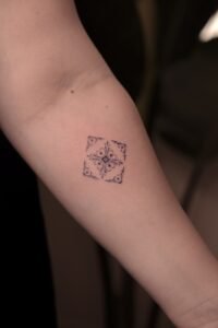 Small geometric tile tattoo with intricate ornamental filigree patterns in a square design, located on the inner forearm