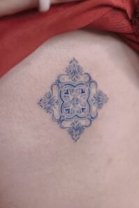 Blue ink ornamental tattoo inspired by azulejo Portuguese tile patterns, featuring a symmetrical diamond-shaped design with intricate fine-line scrolls and floral motifs on the skin