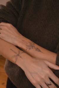 A fine-line Sanskrit script tattoo on the forearm featuring an Om symbol inside a sunburst, a blooming lotus flower, and delicate ornamental swirls, crafted at Names Tattoo Studio in Lisbon