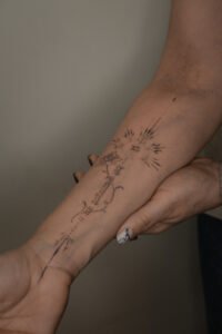 A fine-line Sanskrit script tattoo on the forearm featuring an Om symbol inside a sunburst, a blooming lotus flower, and delicate ornamental swirls, crafted at Names Tattoo Studio in Lisbon