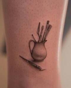 Micro-realistic tattoo of a ceramic pitcher containing various artist paintbrushes and a palette knife on skin