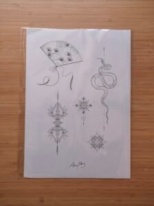 A fine-line tattoo flash sheet featuring an elegant snake, a decorative hand fan