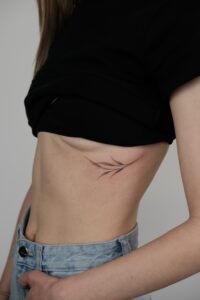 Fine-line minimalist botanical tattoo featuring delicate black ink leaves on a woman's ribs and side torso